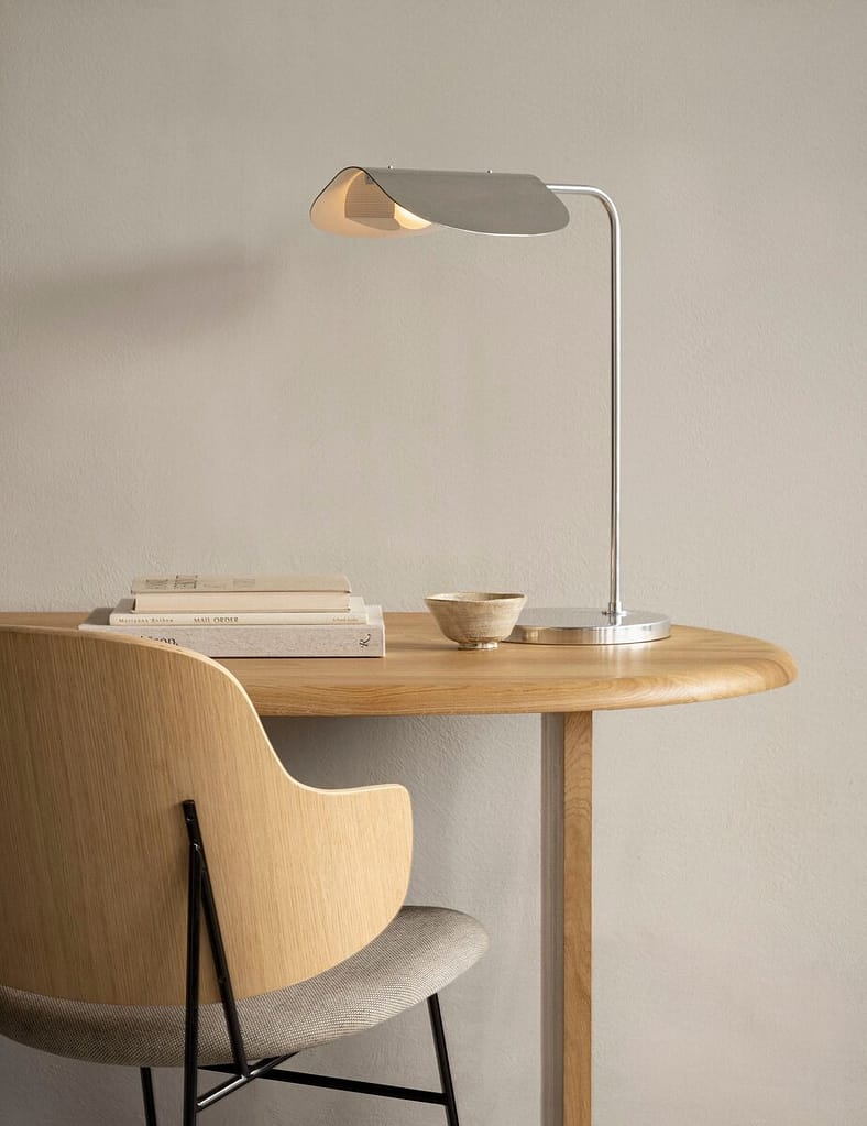 Minimalist Scandinavian-style wooden desk setup with a modern metal desk lamp, a curved wooden chair with fabric seat, a small ceramic bowl, and stacked neutral-toned books against a light beige wall.